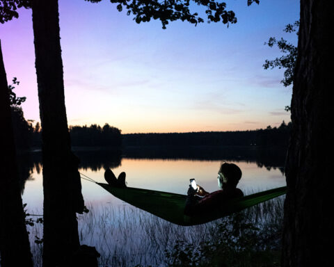 man in hammock near lake with phone in hands late evening