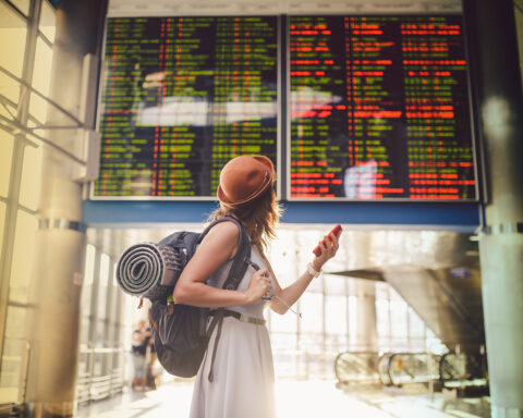Theme travel and tranosport. Beautiful young caucasian woman in dress and backpack standing inside train station or terminal looking at a schedule holding a red phone, uses communication technology