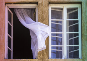 Two windows viewed from outside, one is closed and another is open with white curtain caught on wall stock photo