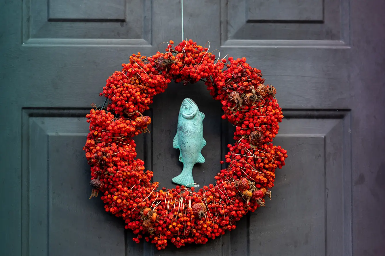 A vibrant red Christmas wreath made of small wild red rowan berries. The decoration is hanging on a black colored door. In the center of the door is a brass door knocker, in the shape of a fish