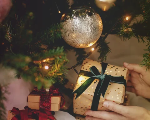 Stylish christmas gift in hands under christmas tree with lights. Merry Christmas and Happy Holidays! Woman in cozy sweater putting wrapped christmas present in atmospheric festive room