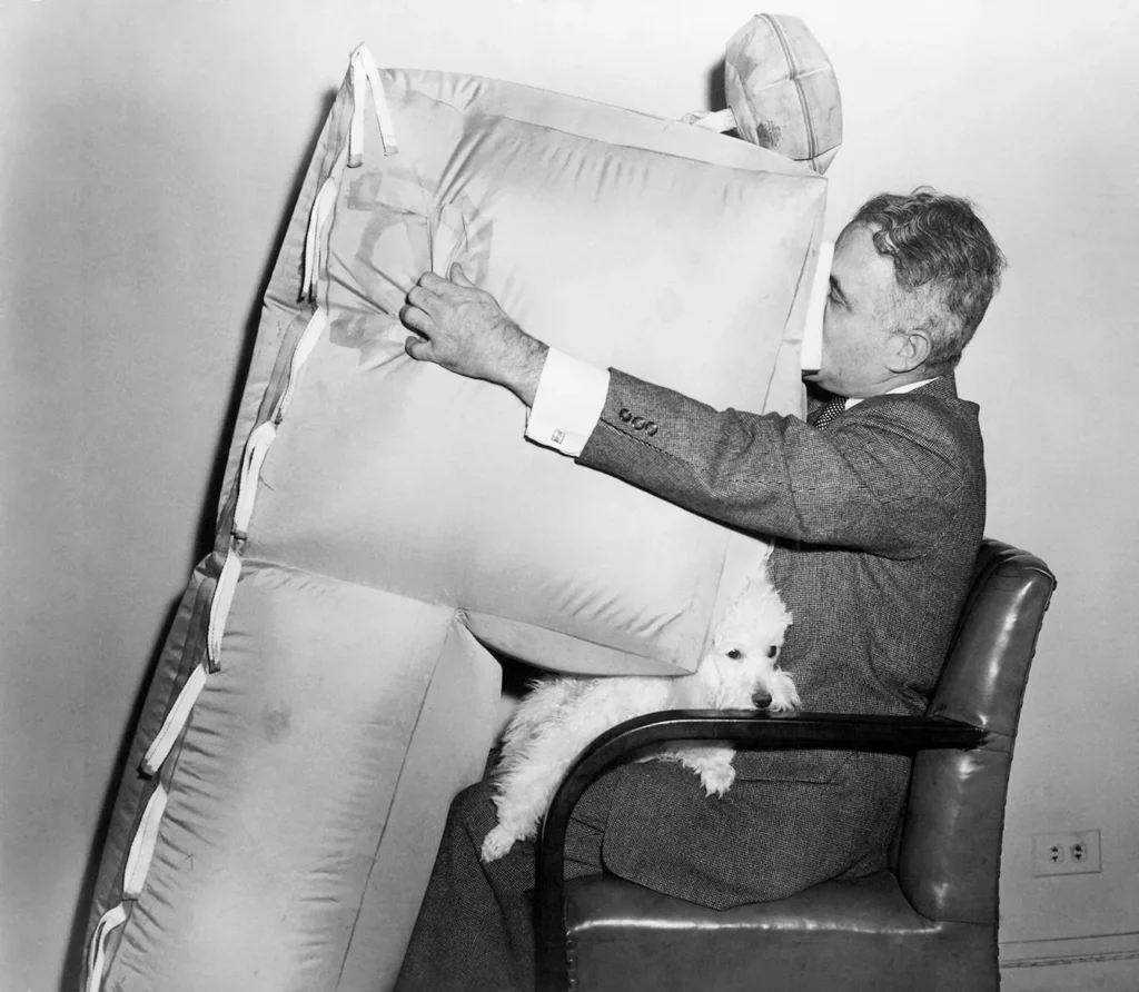 Happier landing for airplane survival crash victims is the purpose of the Micro-Moisture pneumatic safety cushion demonstrated by its designer, Assen Jordanoff, a pioneer aviator, on Jan. 4, 1957. The cushion, which is being studied by military and civil aviation authorities, is installed in the back of airplane seats. By throwing a single switch, the pilot can inflate a planeful in three seconds. The French poodle shows how a child on a parent's lap would be protected from the crushing jolt of a crash landing or ditching