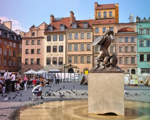 Warsaw, Poland. Main Square of the Old Town. Warsaw Mermaid - symbol of the city - in front on the right side, many tourists looking visiting the place and feeding pigeons on the left side, and the colorful antique tenements in the background.