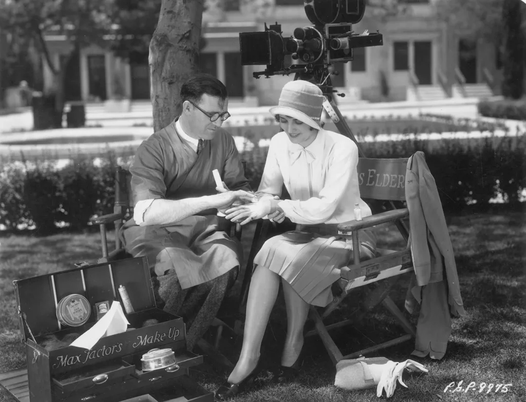 1928: American aviator and actress Ruth Elder (1902 - 1977) sits in a folding chair while makeup man Jim Collins smoothes foundation into her hand outdoors on the set of director Frank R. Strayer's silent film, 'Moran of the Marines'. A Max Factor make-up case rests at Collins' feet.