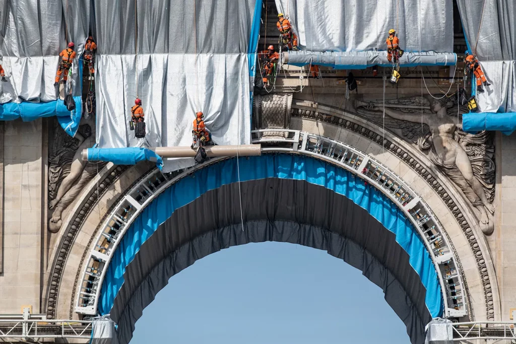 Arc De Triomphe To Be Wrapped For Posthumous Work By Artist Christo. Workers begin the process of wrapping up the Arc De Triomphe monument in silver-blue fabric on September 12, 2021, in Paris, France. The monumental installation will wrap the landmark Parisian monument under a 25,000 square meters silver and blue fabric. "L' Arc de Triomphe, Wrapped" project by late artist Christo and Jeanne-Claude will be on view from September 18 to October 03, 2021. Bulgarian-born US artist Christo died in May 2020.