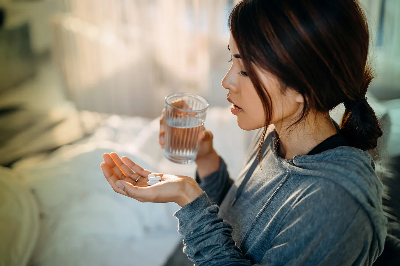 Young woman sitting on bed and feeling sick, taking medicines in hand with a glass of water, stock photo.