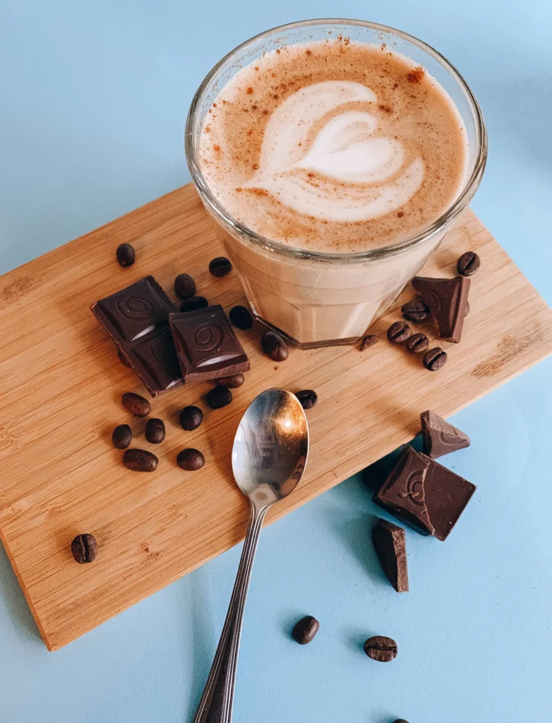 The coffee drink in a glass stands on a board that is strewn with coffee beans and chocolate.