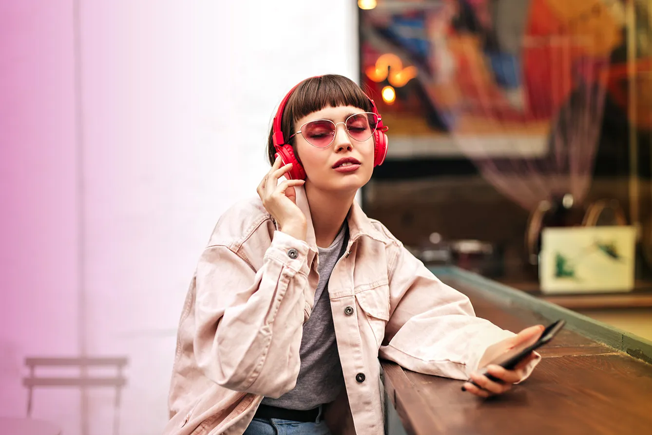 Trendy woman with short hair in headphones listening to music outdoors. Beautiful girl in cool denim jacket and bright glasses holding phone