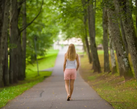 Young adult blonde woman slowly walking through alley of green trees on sidewalk at city park in warm summer day.