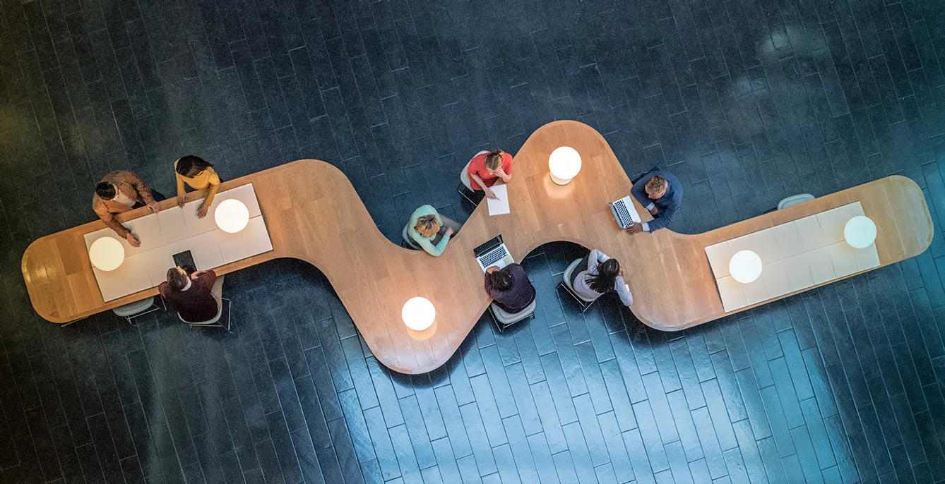 Panoramic overhead view of several business meetings going on in the communal area of a modern office building.