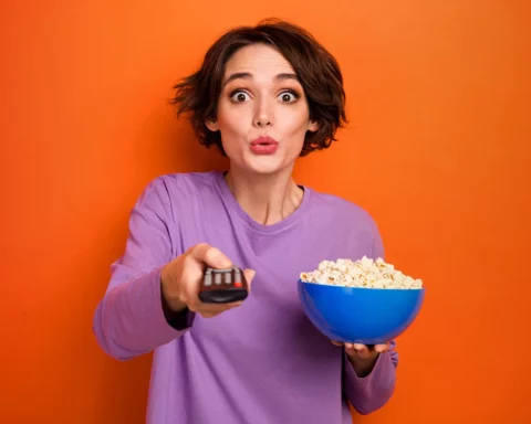 Portrait of impressed nice girl hold popcorn plate switch tv program isolated on orange color background.