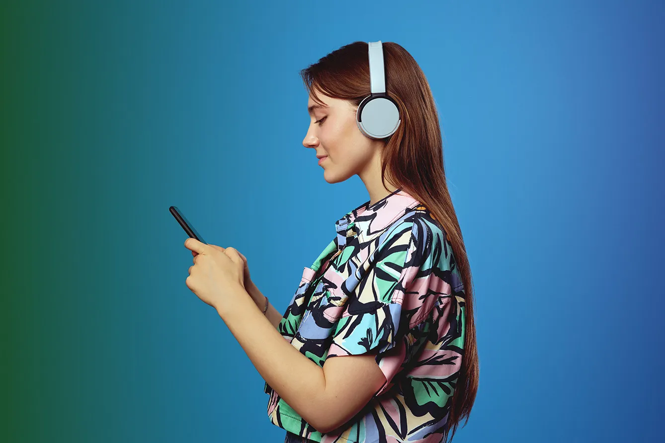 Side view of young student girl using mobile phone and looking at screen, wearing headphones while listening to music.